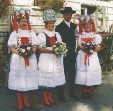 Drei Frauen in Festtracht mit Brautkronen neben einem Mann in dunklem Anzug und Hut